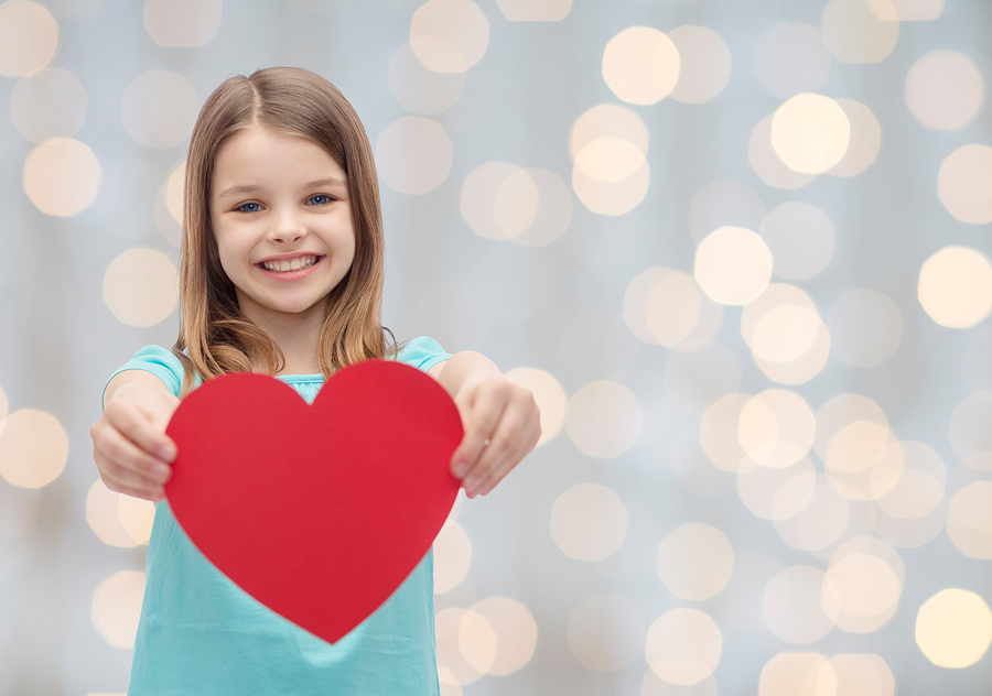 love, charity, holidays, children and people concept - smiling little girl with red heart over lights background